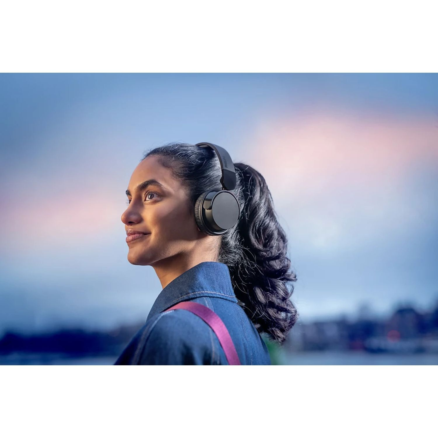 Woman with headphones looks away. Blue sky background. Pink strap. Denim jacket. Ponytail.