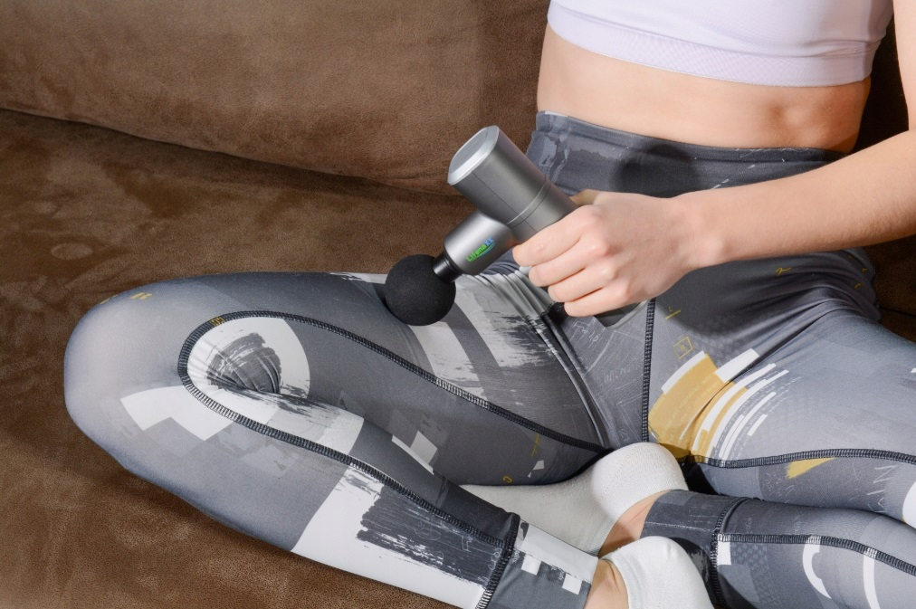 Kobieta używająca pistoletu do masażu na udzie siedząc na kanapie. Ubrana w strój sportowy.