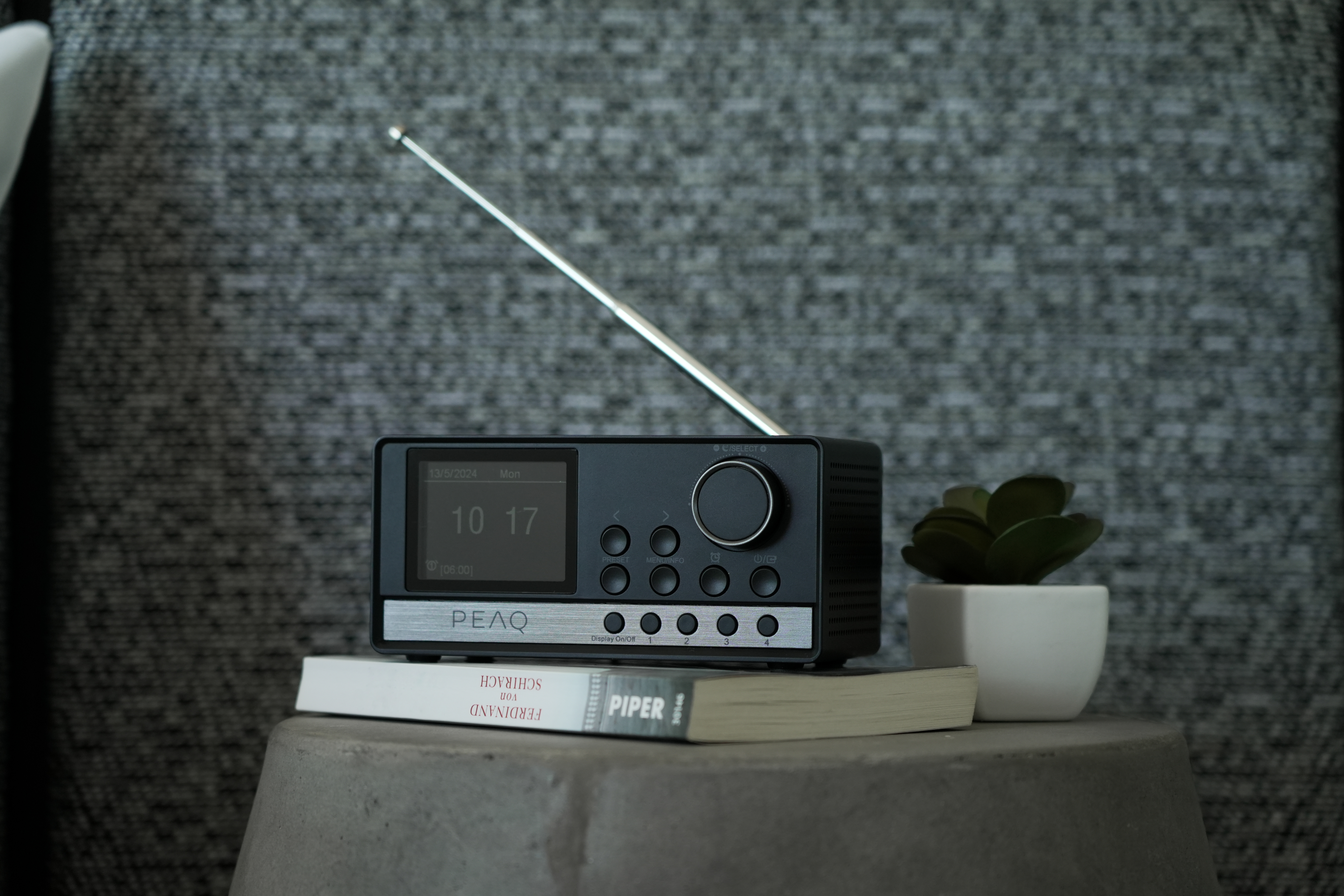 Ein schwarz-silbernes Radio, ein Buch und eine kleine Pflanze auf einer grauen Oberfläche.