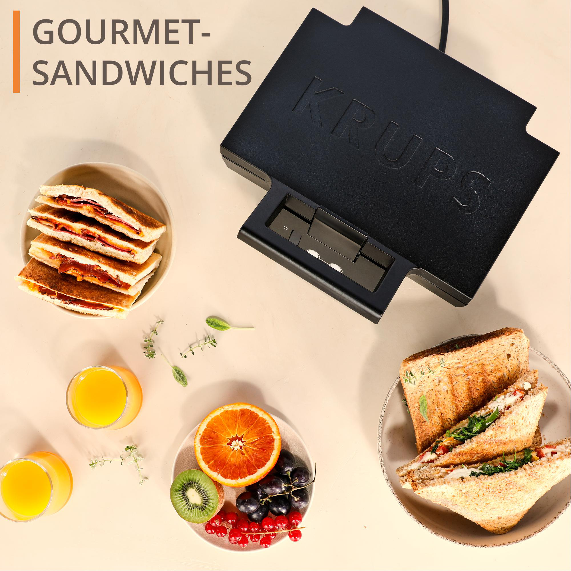 Sandwiches, Orangensaft und Obst neben einem Krups Sandwichmaker. Weißer Hintergrund.