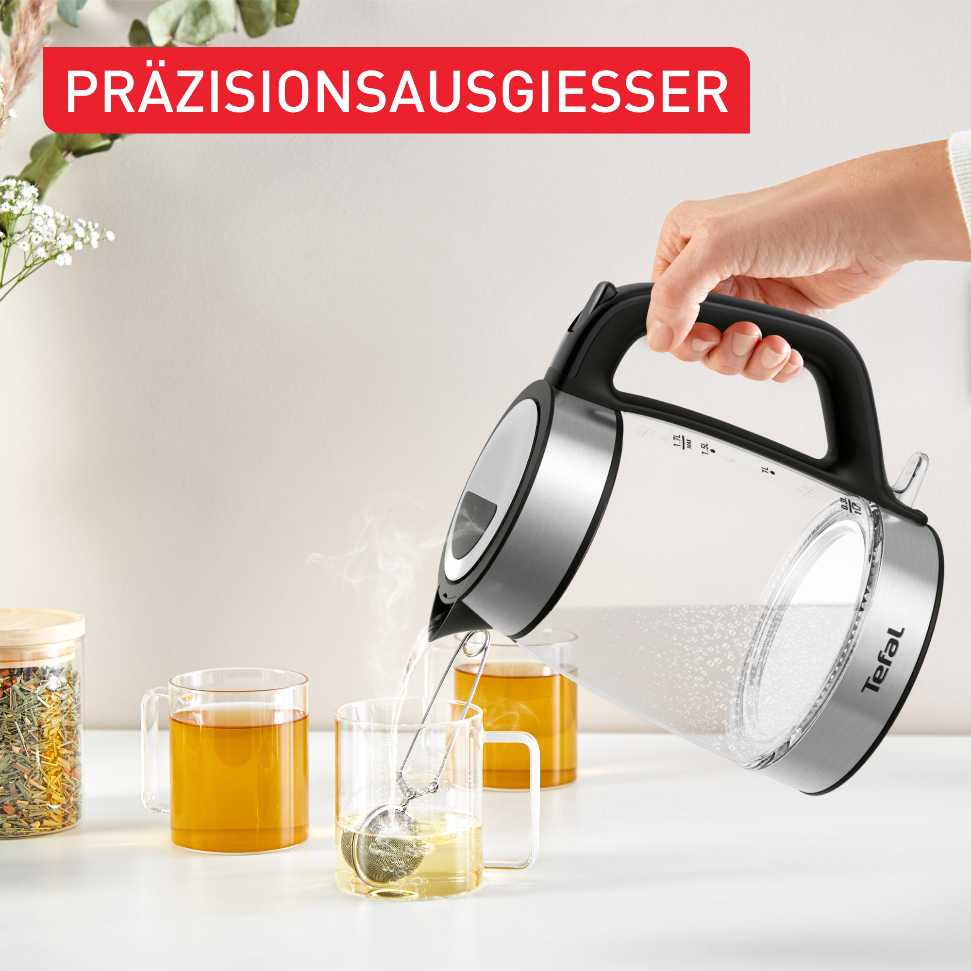 Eine Person gießt heißes Wasser aus einem elektrischen Glaskessel in eine Tasse mit Tee-Infuser.