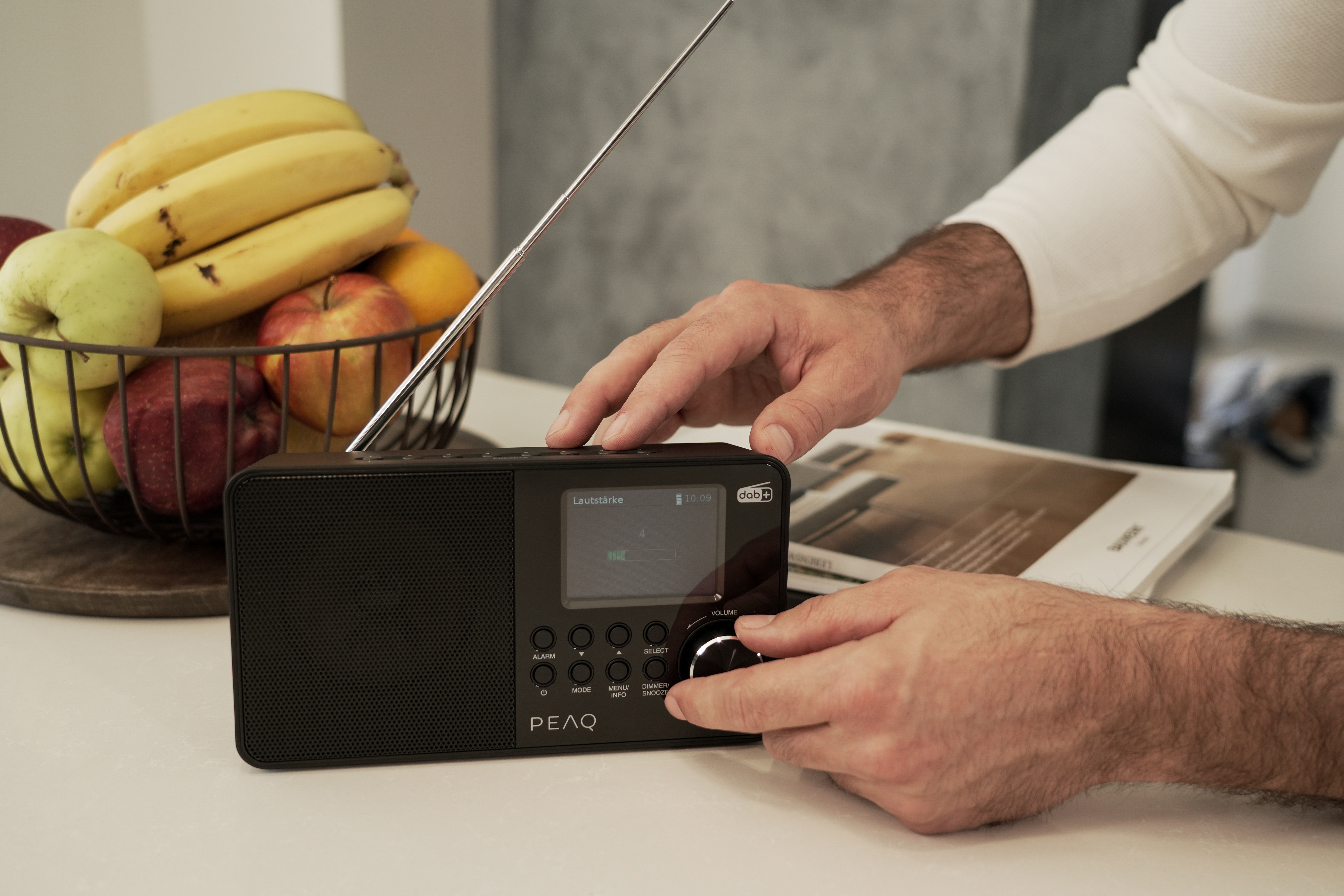 Hände eines Mannes, die den Lautstärkeregler an einem schwarzen Radio einstellen, mit Obst davor.