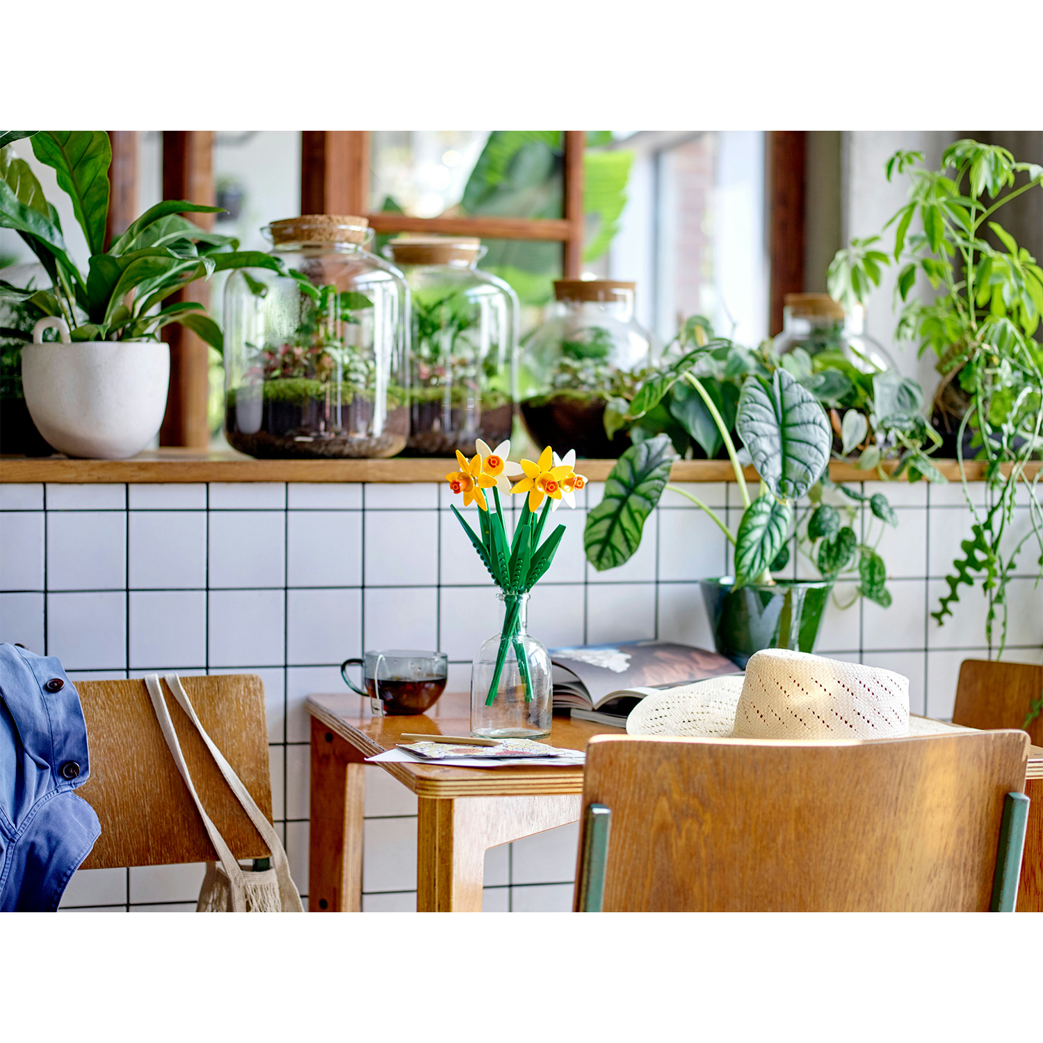 Scena interna con piante, fiori e un tavolino. Include vasi di vetro e un muro piastrellato bianco.