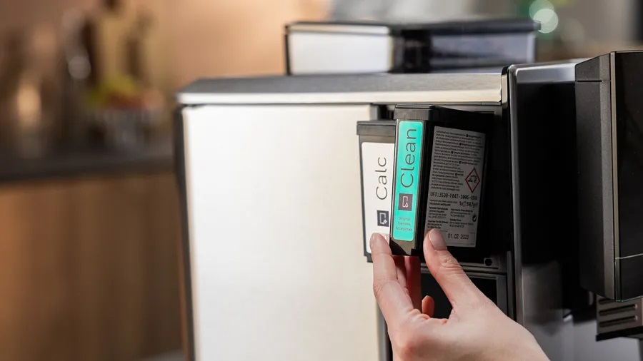 Ręka wkładająca wkład 'Clean' do ekspresu do kawy, obok wkład 'Calc'.