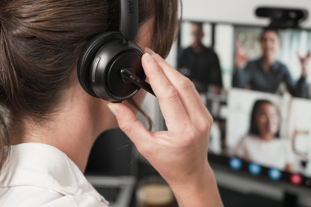 Woman wearing headphones on a video call. Other people are visible on a screen in the background.
