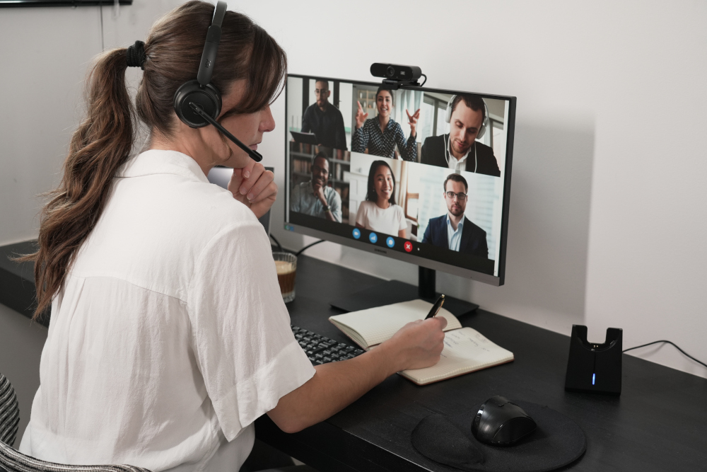 A woman in a white shirt is in a video call on a monitor, taking notes and wearing a headset.