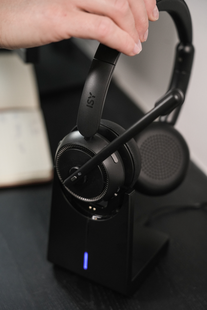 A black headset on a black charging stand, held by a hand. The stand has a blue light.