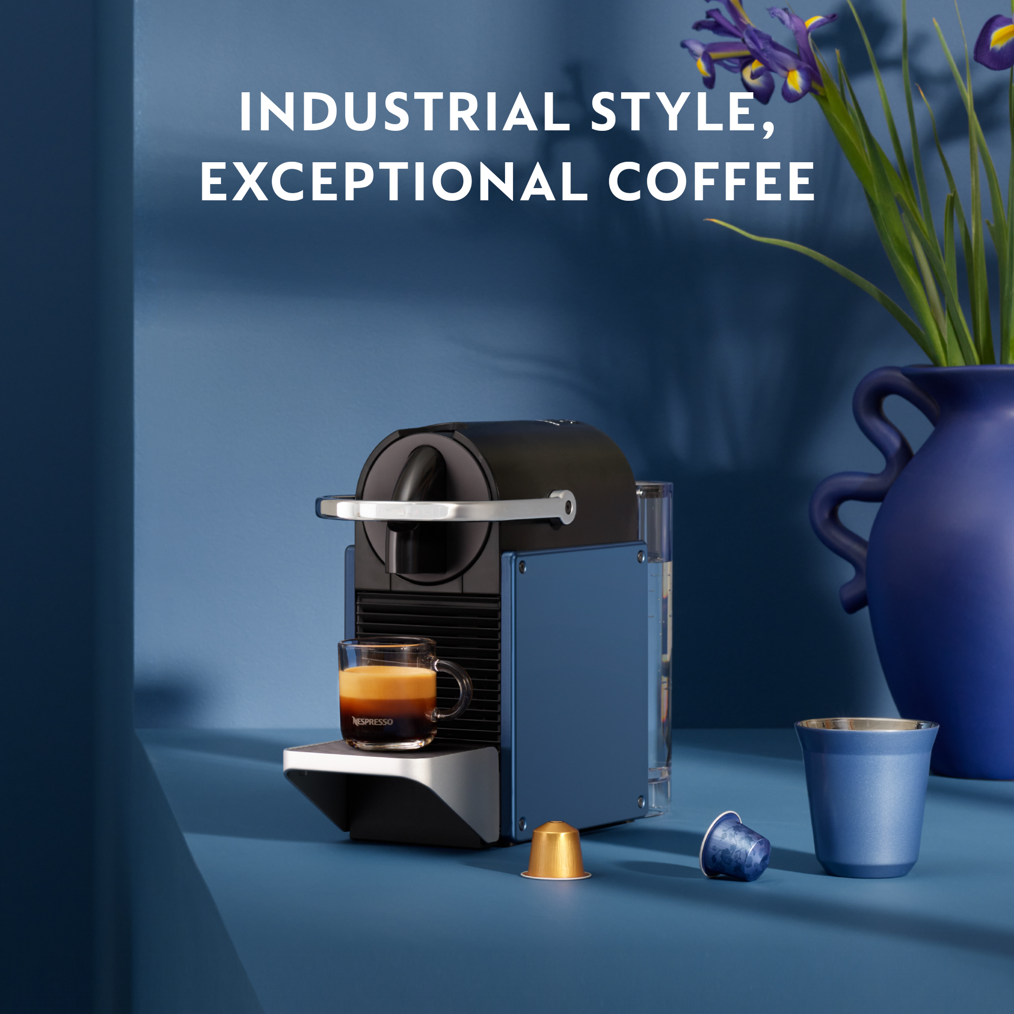 Blaue und schwarze Espressomaschine mit einem Glas Kaffee, Kaffeekapseln und einer blauen Tasse auf einem Tisch.