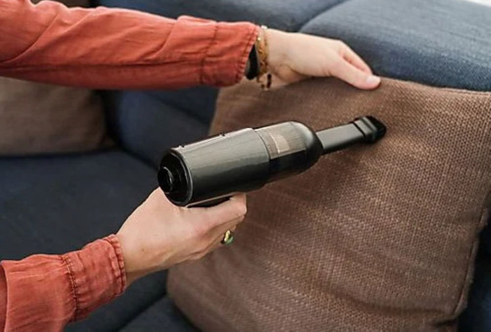 Osoba odkurza brązową poduszkę na niebieskiej sofie czarnym odkurzaczem ręcznym.