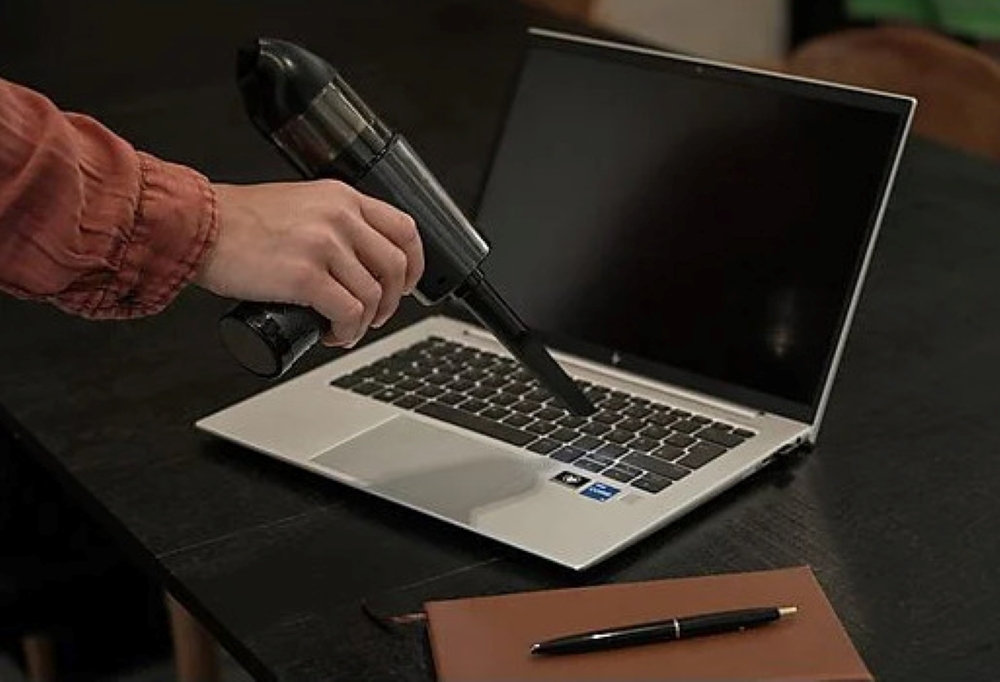 Osoba używająca czarnego odkurzacza ręcznego do czyszczenia klawiatury laptopa na biurku.