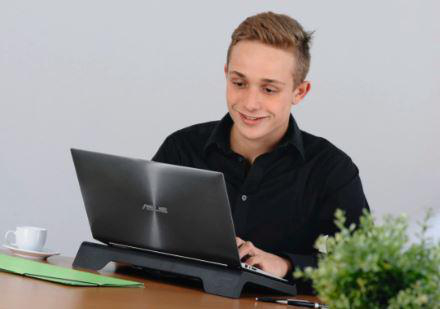 Ein junger Mann lächelt, während er an einem Laptop arbeitet, mit einer Kaffeetasse und Pflanze.