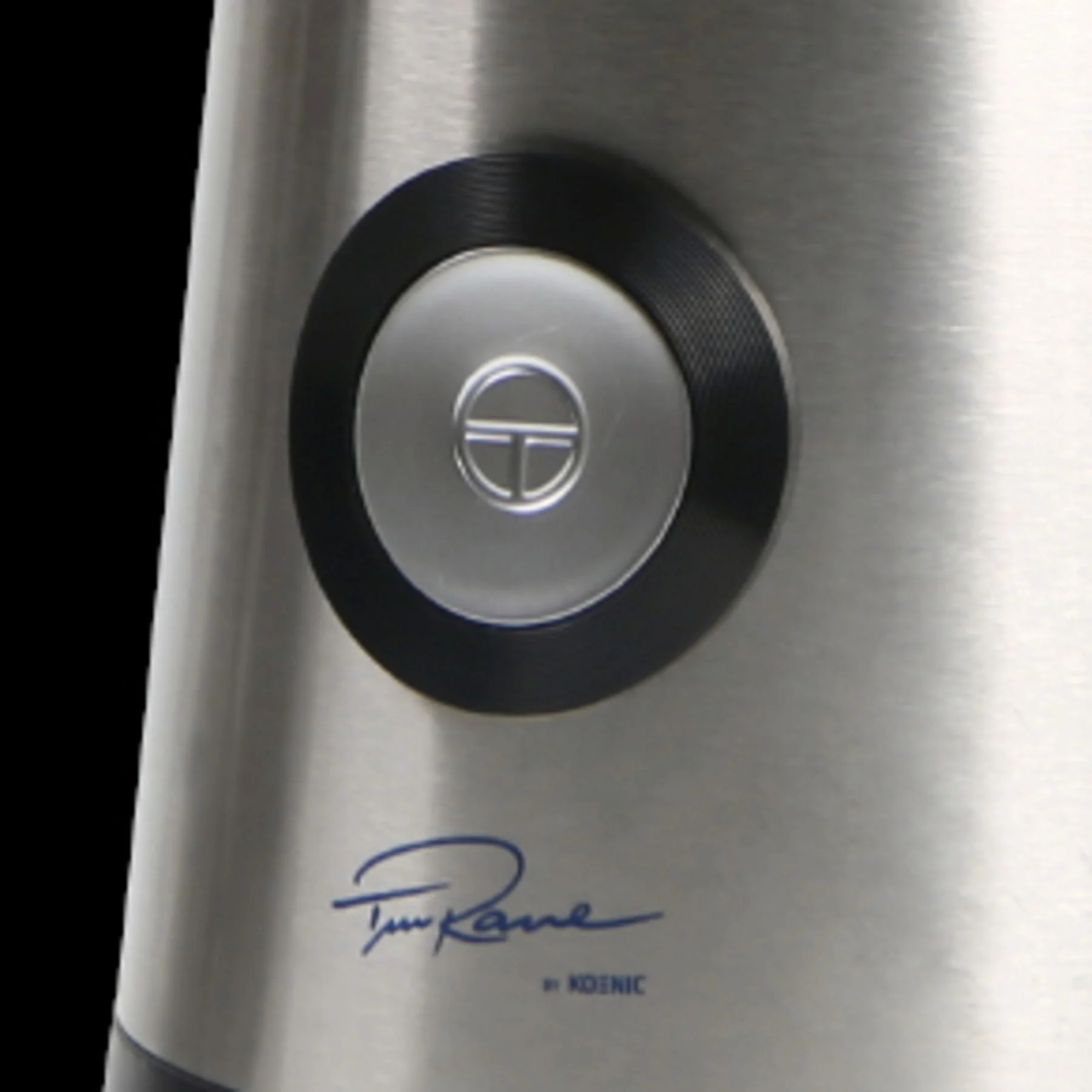 Zbliżenie srebrnego przycisku z czarnym pierścieniem, z wygrawerowanym 'T' w okręgu.