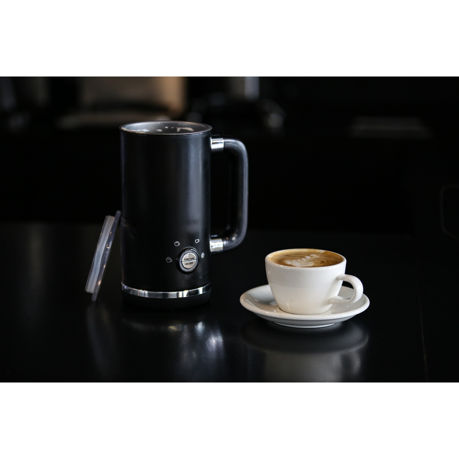Zwarte melkopschuimer en witte kop koffie op een zwart oppervlak. Een deksel ligt naast de opschuimer.