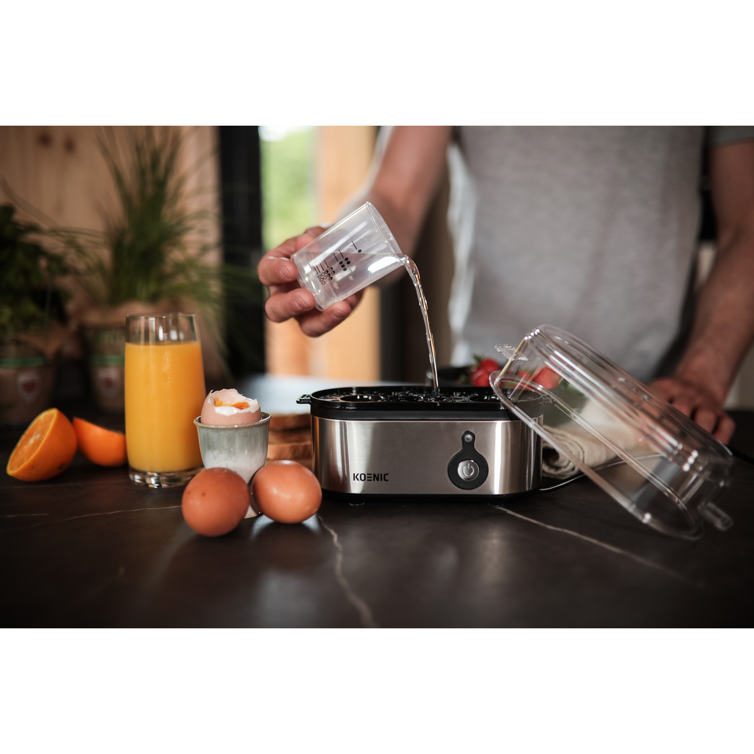 A person pouring water into an egg boiler. Orange juice, eggs, and fruit are visible.