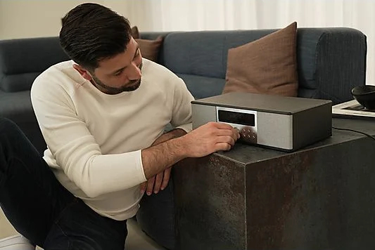 Mężczyzna siedzący na podłodze reguluje głośność w zestawie stereo z srebrnym panelem przednim.