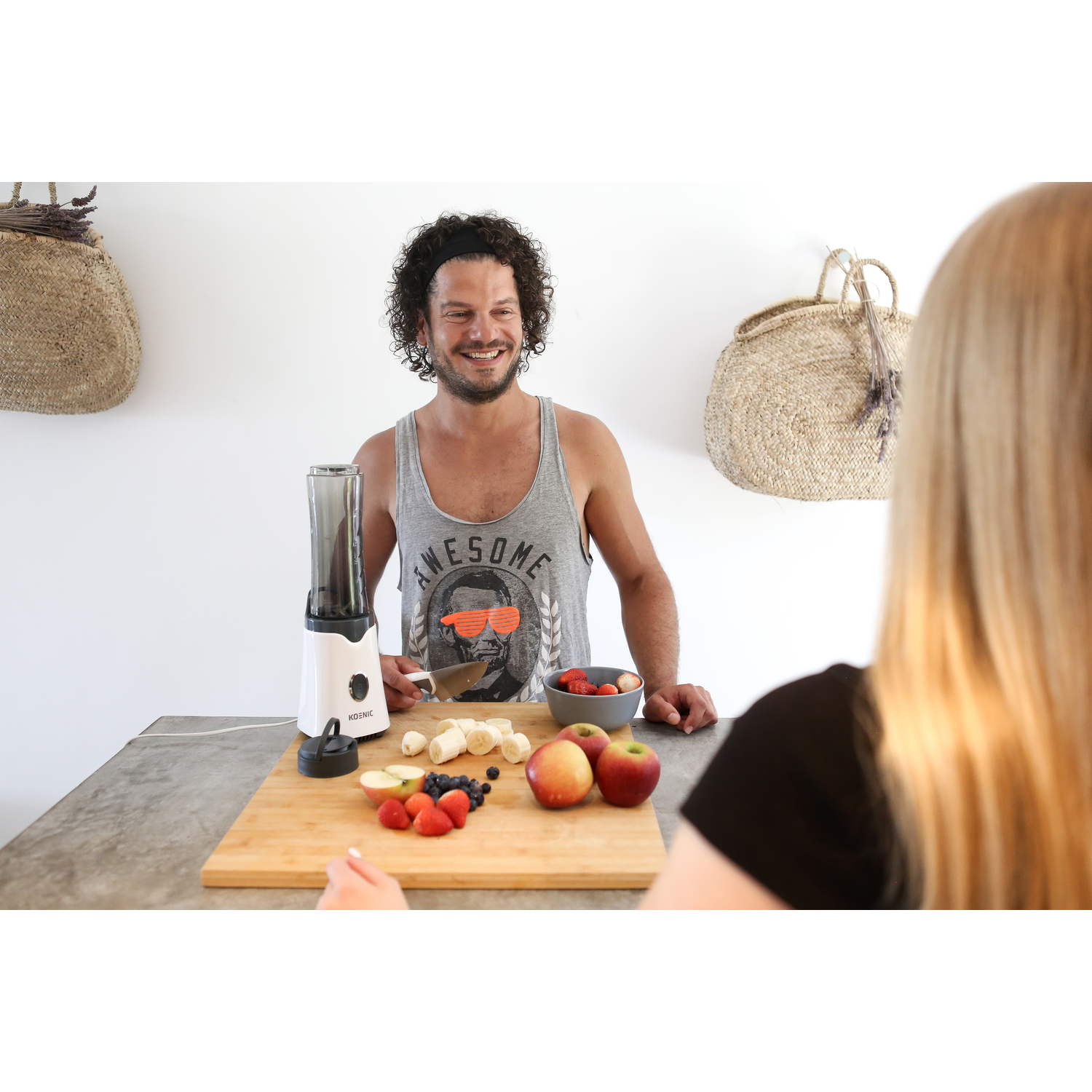 A man is preparing fruit with a blender on a table, another person is watching him.