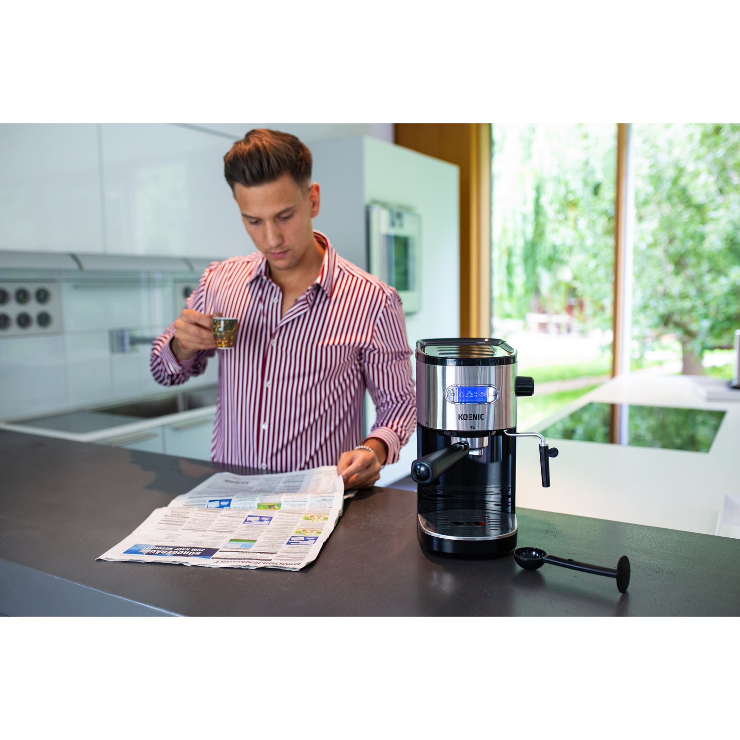 Man in keuken, drinkt uit een kopje, leest een krant. Espressomachine op de achtergrond.