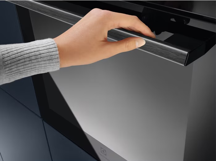 Ręka otwierająca czarny uchwyt drzwi piekarnika, z rozmytym tłem.