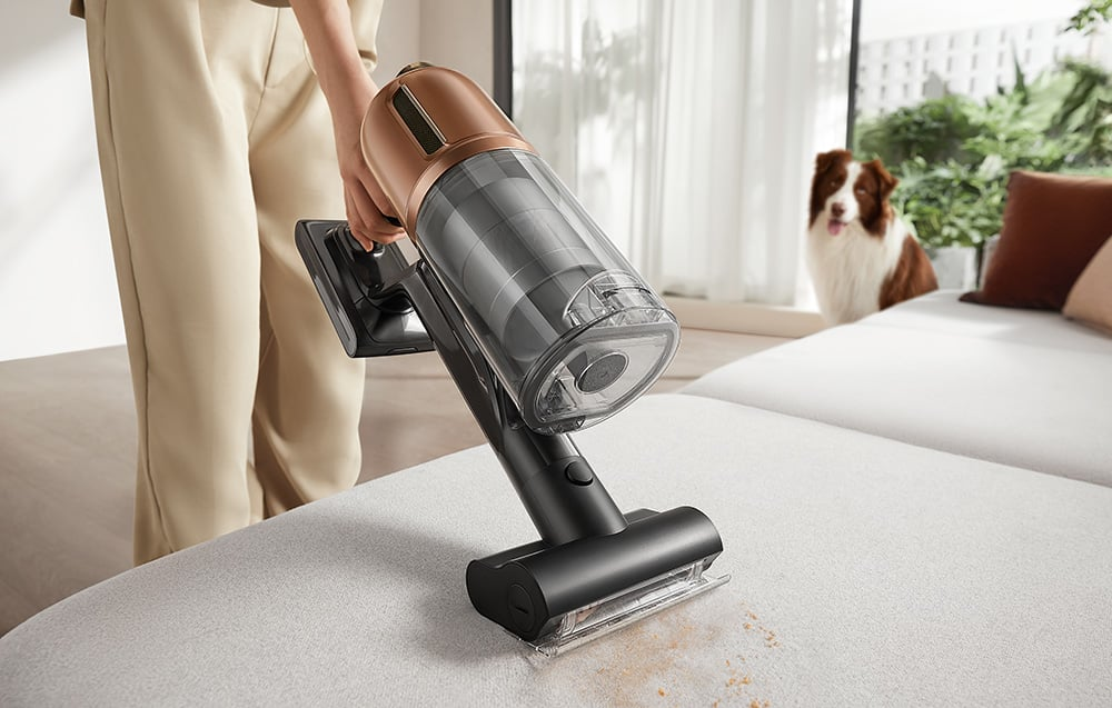 Osoba używająca odkurzacza bezprzewodowego na kanapie, pies w tle.