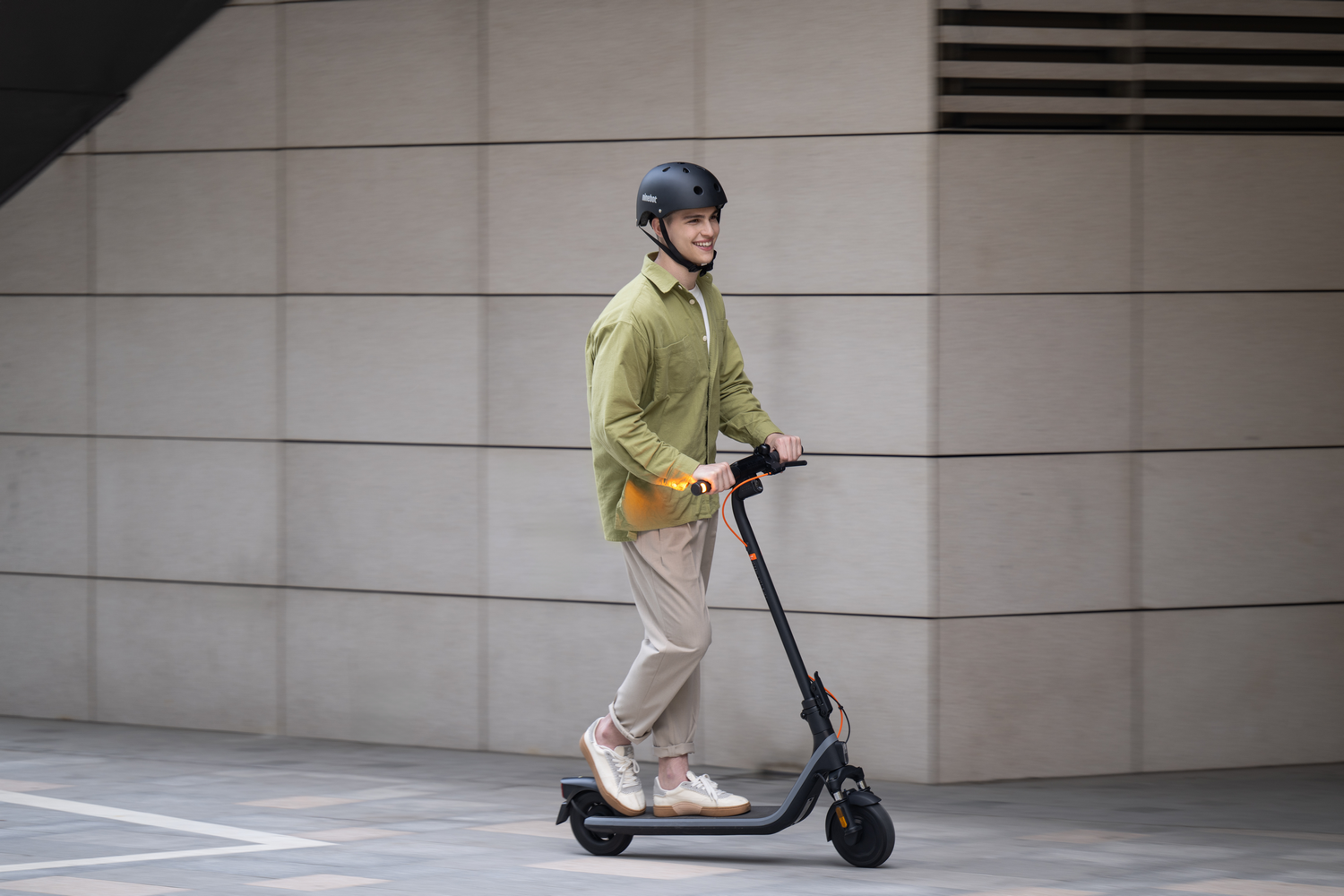 Un hombre con ropa casual monta un patinete eléctrico negro y naranja en un camino pavimentado.