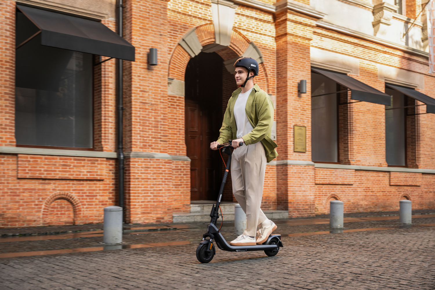 Un hombre monta un patinete eléctrico en una calle adoquinada frente a un edificio de ladrillo.