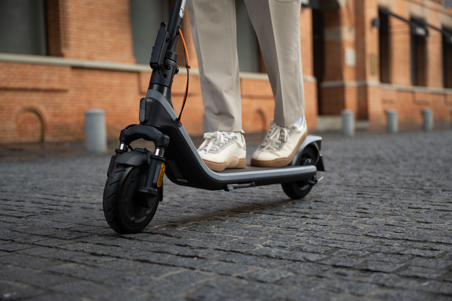 Una persona montando un patinete eléctrico negro y gris en una calle adoquinada, con pantalones y zapatos color beige.