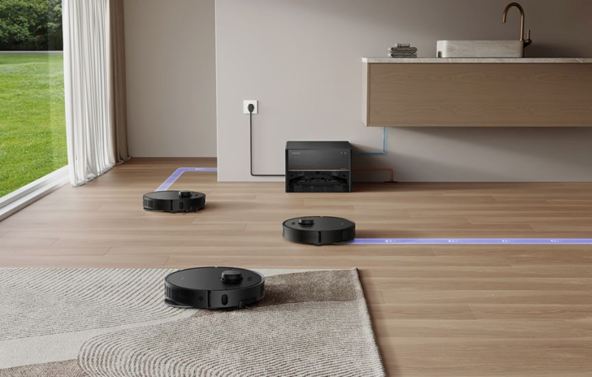 Trzy czarne roboty sprzątające na drewnianej podłodze, jeden na dywanie. Stacja ładująca pośrodku.