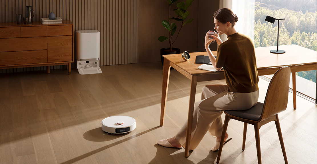 Une femme dans une pièce avec un aspirateur robot. Elle est à une table en train de boire dans une tasse.