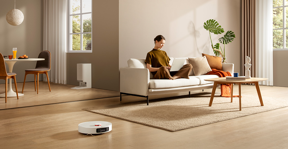 Une femme utilise un ordinateur portable sur un canapé blanc. Un aspirateur robot est sur le sol en bois.