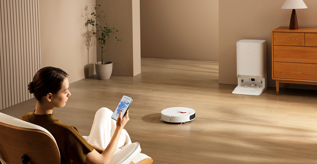 Une femme utilise un téléphone pour contrôler un aspirateur robot. L'aspirateur est au sol. Il y a une station.