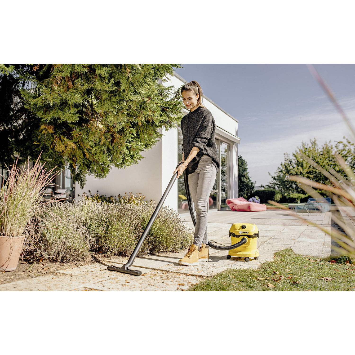 A woman vacuuming a patio with a yellow vacuum cleaner, plants, and a house in the background.