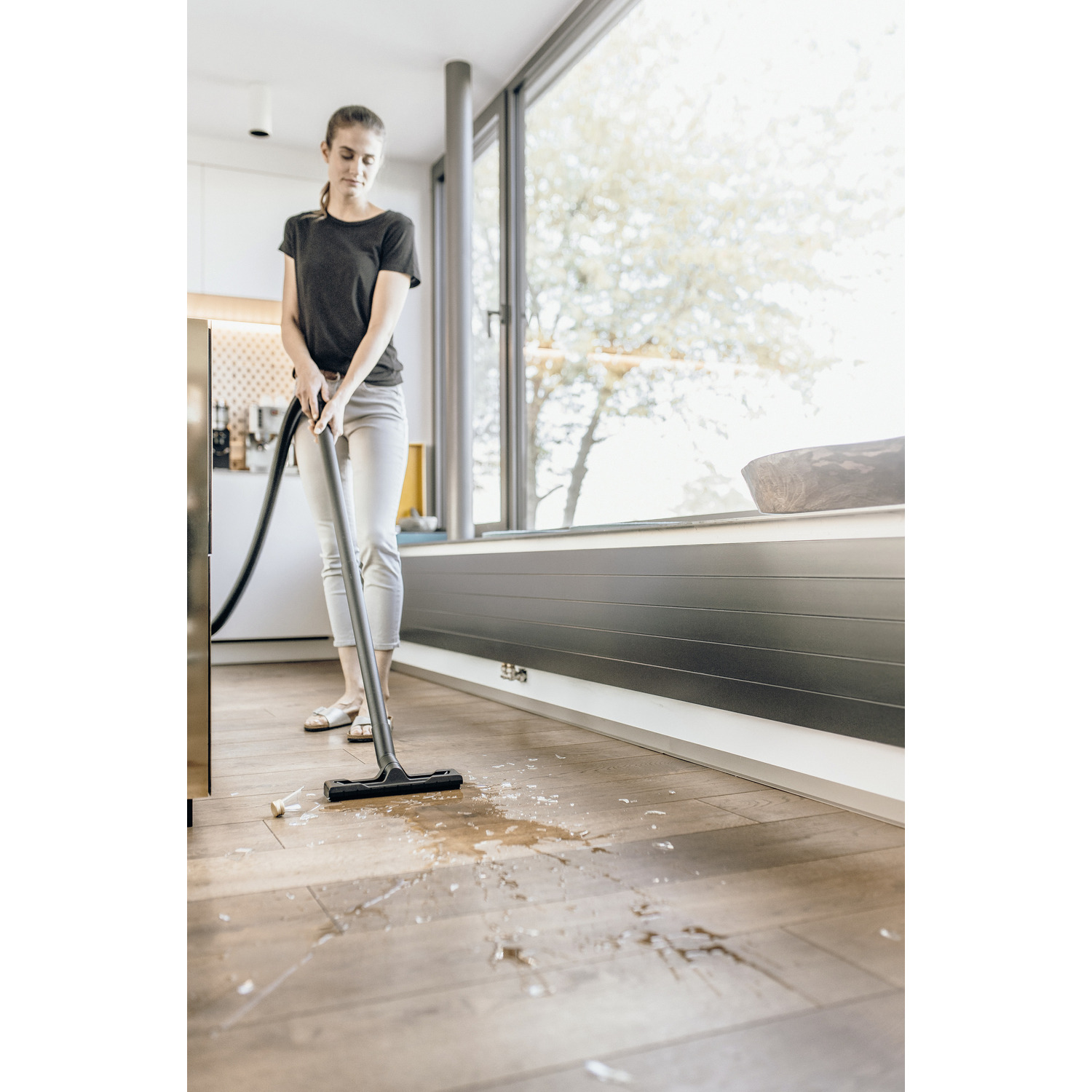 A woman vacuuming a wooden floor with water on it. She is in a kitchen.