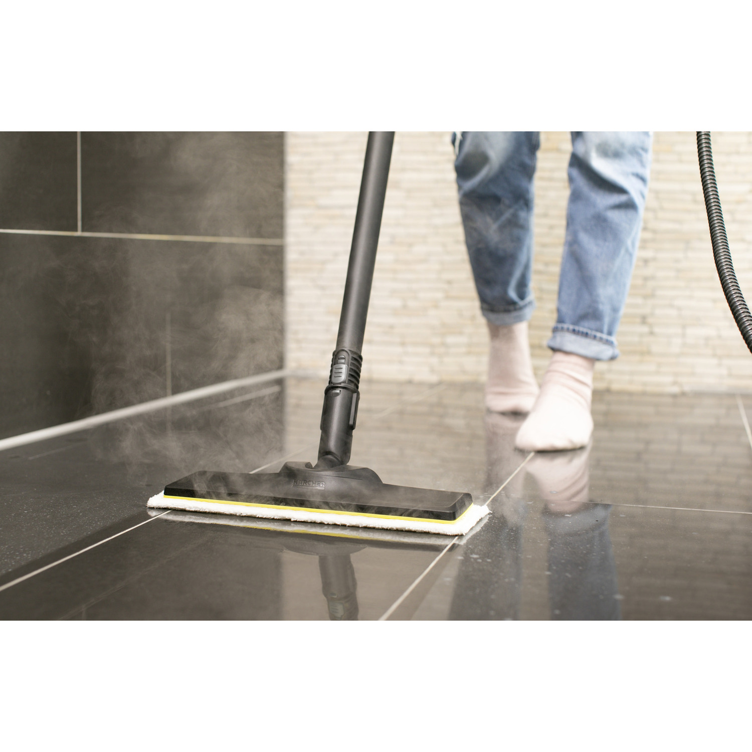 A person, wearing jeans and socks, using a steam cleaner on a tiled floor, creating steam.