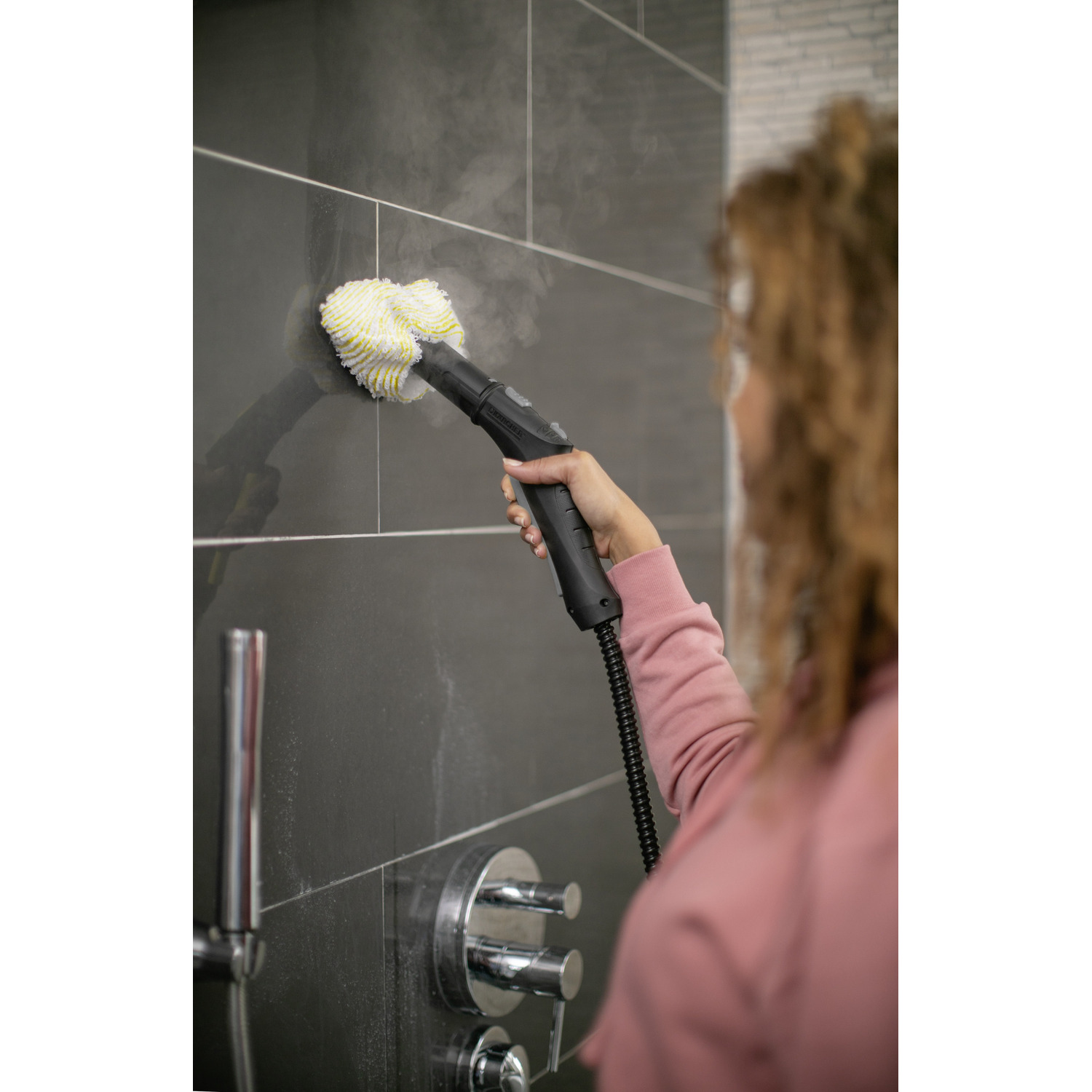 A woman using a steam cleaner to clean a tiled shower wall. Steam is visible.