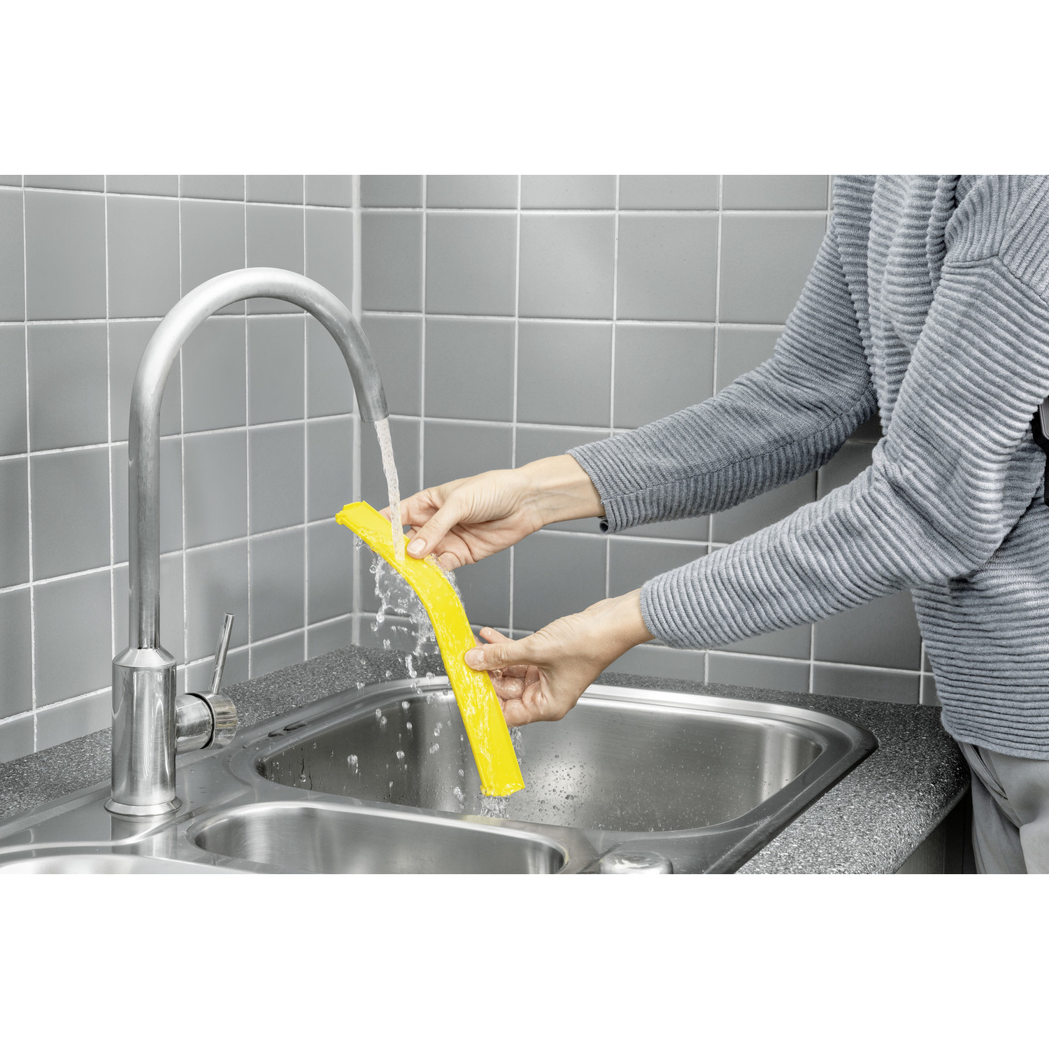De handen van een persoon houden een gele schoonmaaktool onder stromend water in een gootsteen.
