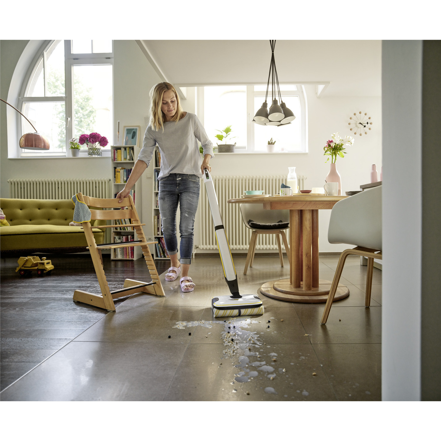 Woman cleans floor with a white vacuum. A wooden high chair and a yellow toy truck are in the background.