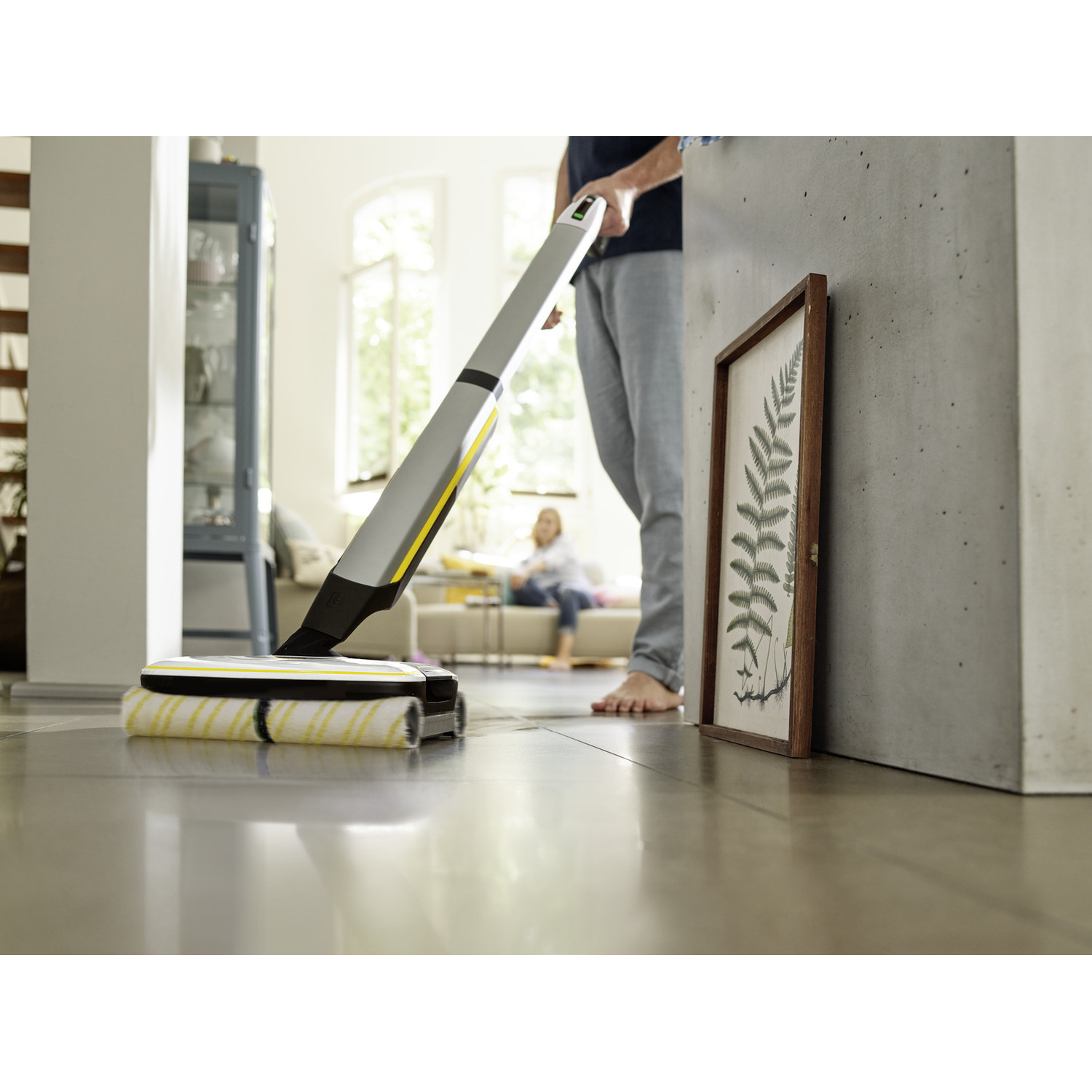 A person using a mop, a wooden framed picture on the right, and a person sitting on the couch.