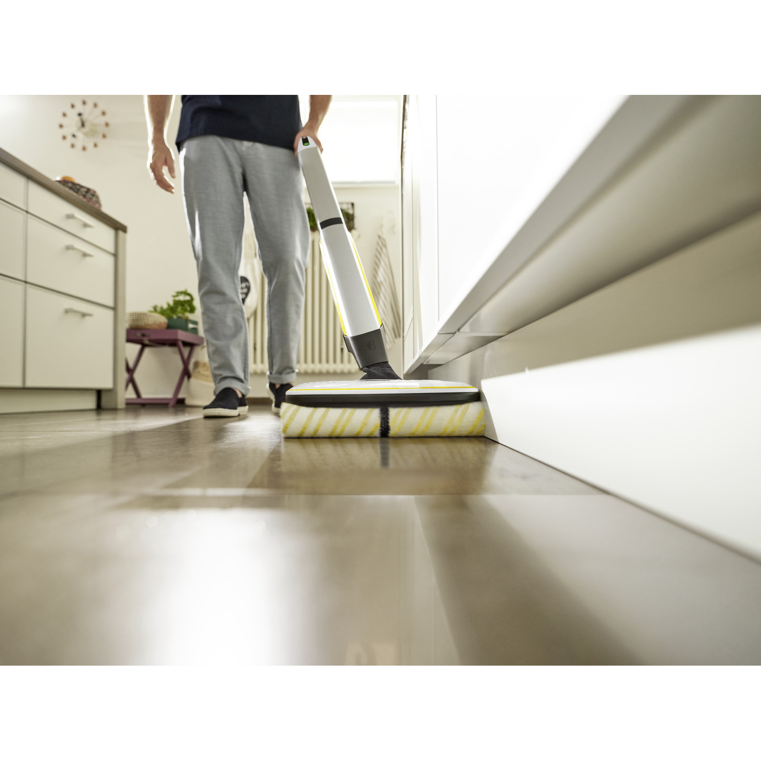 A person vacuuming a wooden floor with a yellow and white cleaning device, near a white wall.