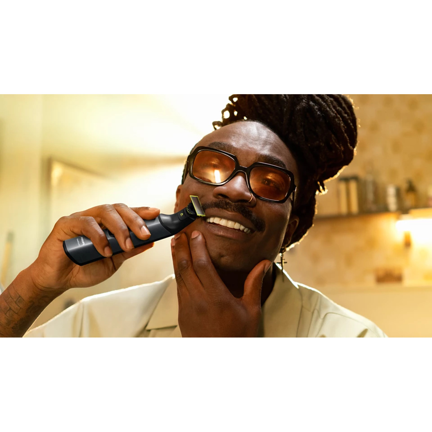 A man shaves his cheek with a black trimmer. He wears glasses and has dreadlocks, smiling at the camera.