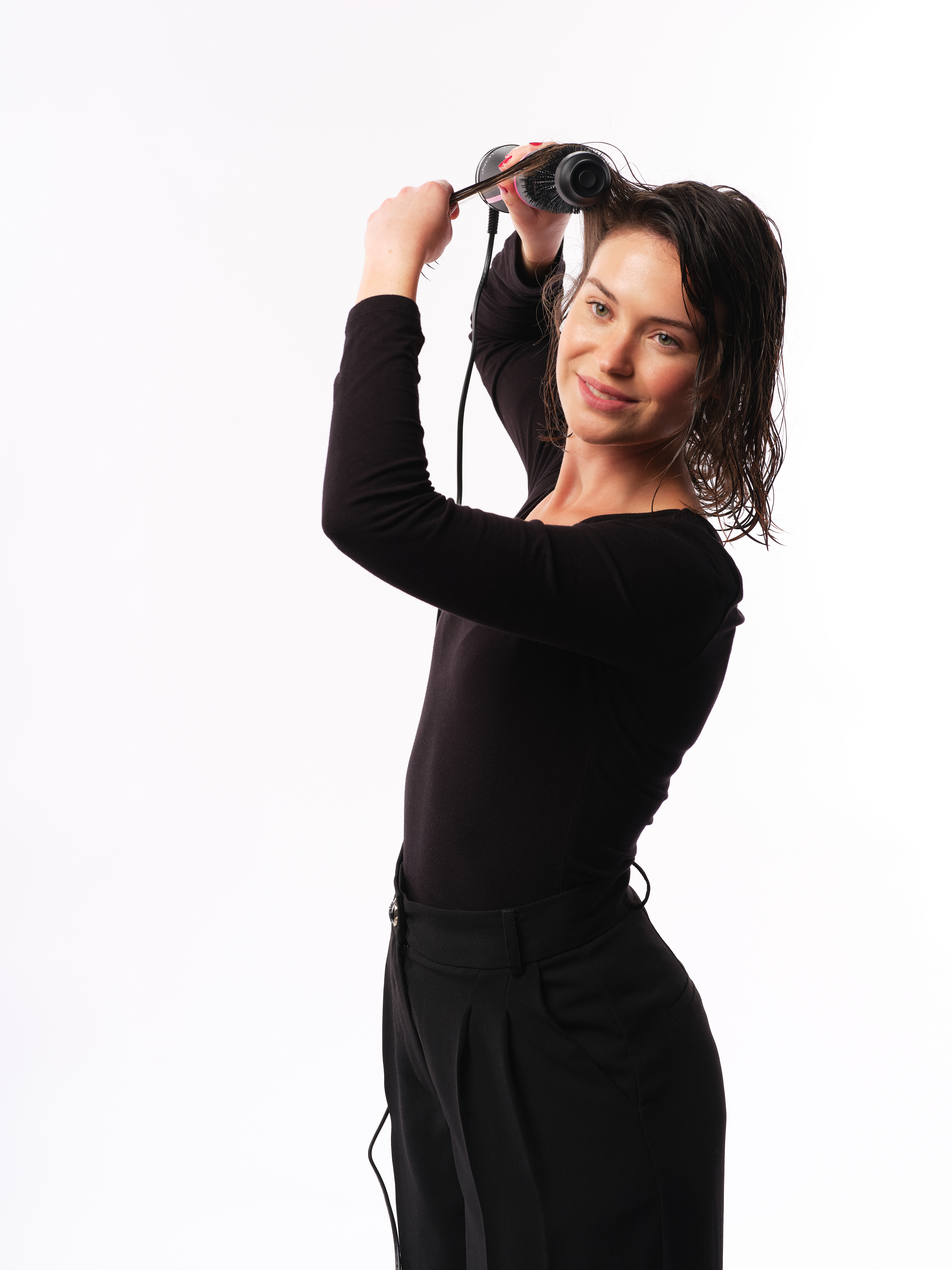 Frau trocknet ihr Haar mit einem Haarstyler, lächelt in die Kamera. Weißer Hintergrund.