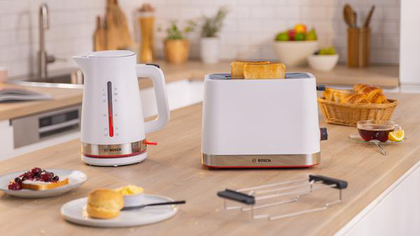 Ein weißer Wasserkocher und Toaster mit Brot, Butter und anderen Gegenständen auf einer Holzplatte.