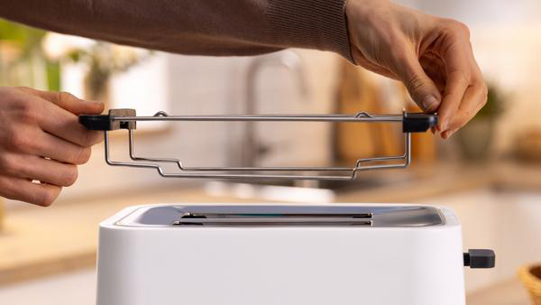 Hände, die ein Metallgestell in einen weißen Toaster einsetzen, Küchenhintergrund sichtbar.