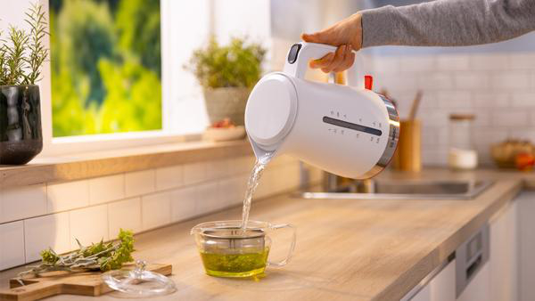 Eine Hand gießt Wasser aus einem weißen Wasserkocher in eine Tasse.