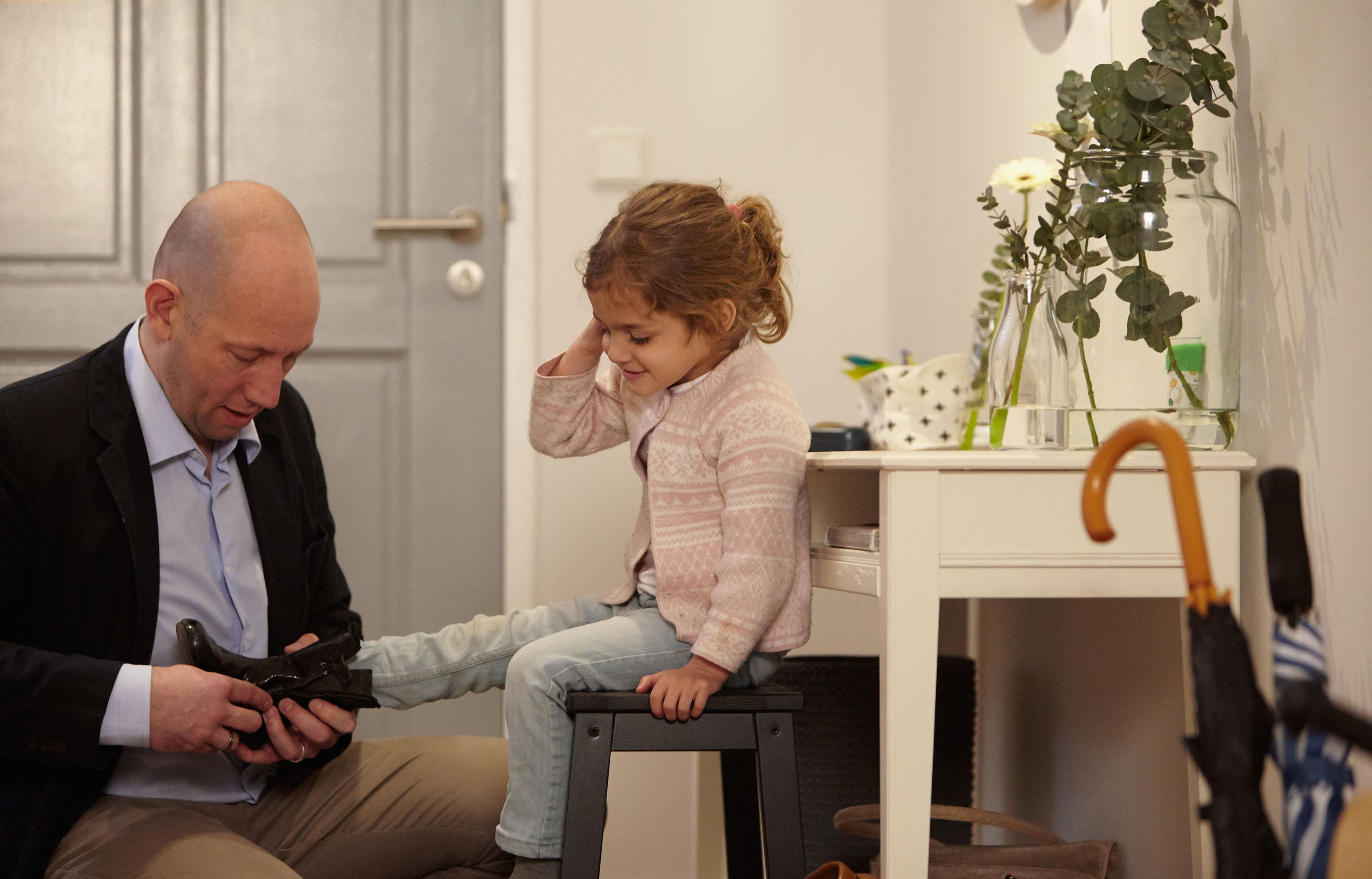 Ein Mann zieht einem Kind einen Schuh an. Das Kind sitzt auf einem Hocker und lächelt.