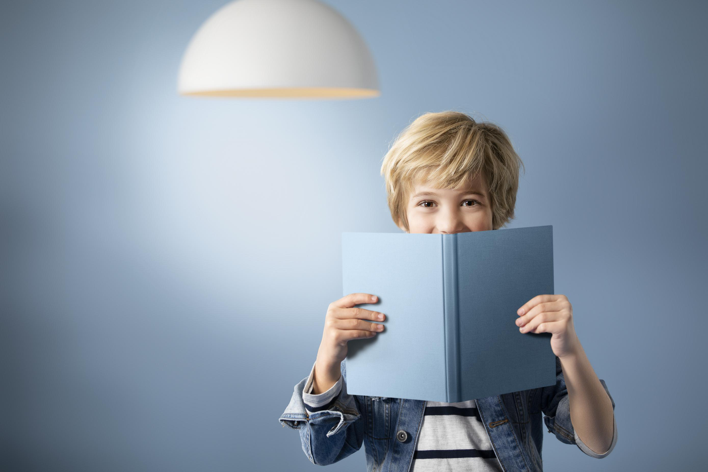 Ein Junge mit blonden Haaren hält ein blaues Buch vor sein Gesicht und schaut darüber. Über ihm ist ein Licht.