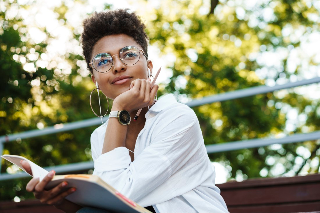 Femme en lunettes, chemise blanche, cahier, écouteurs dans un parc.