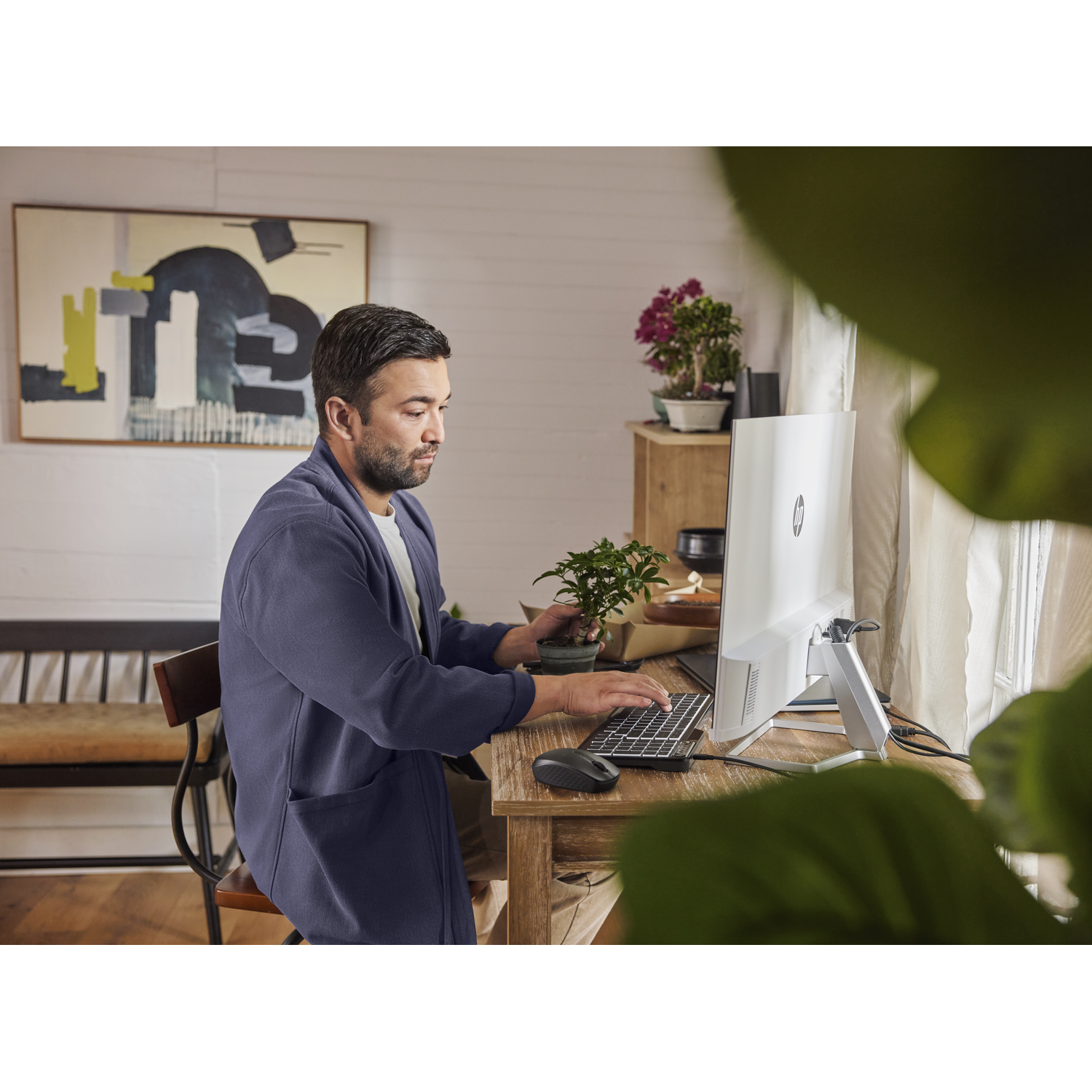 Man sitting at a desk with a computer, holding a plant. A painting and other items are in the background.