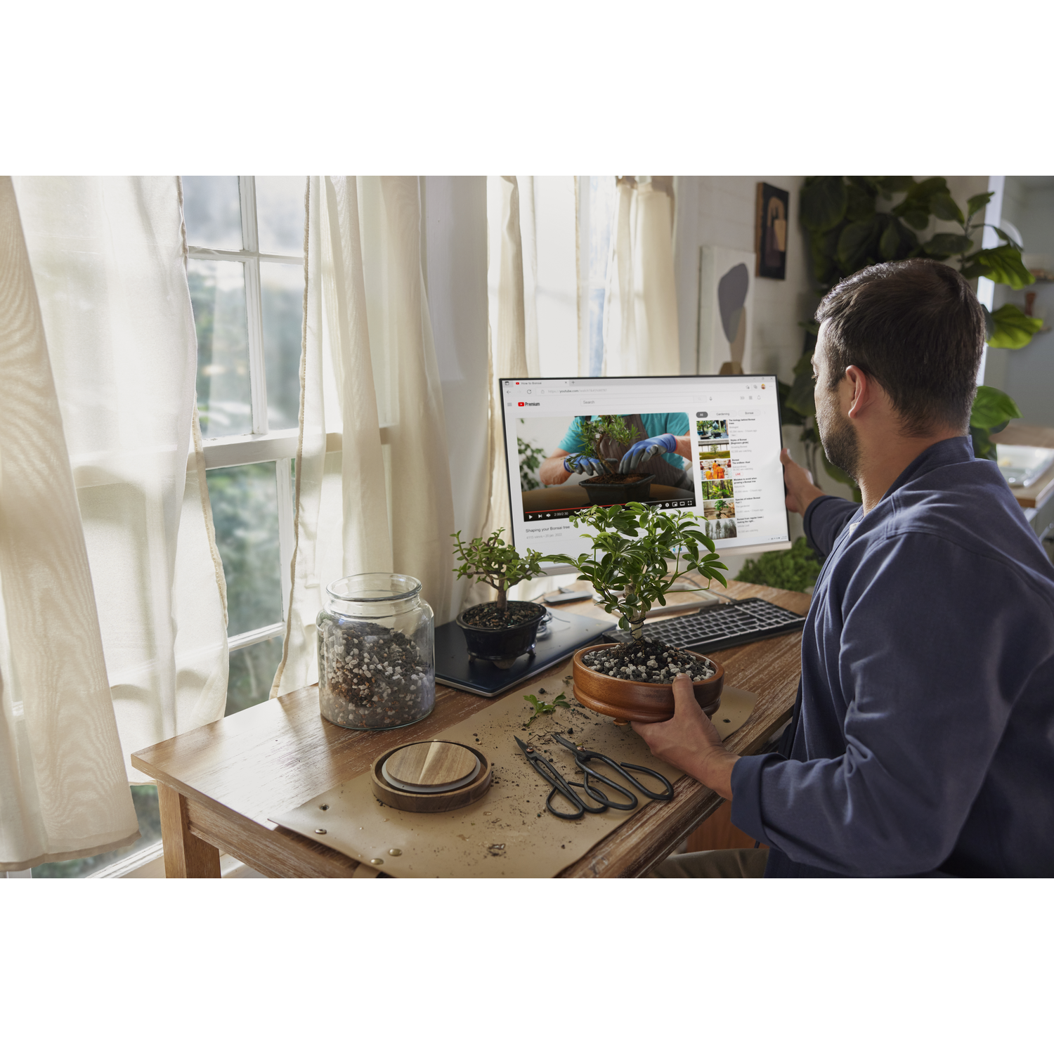 A man tending bonsai trees, watching a tutorial on a computer screen.