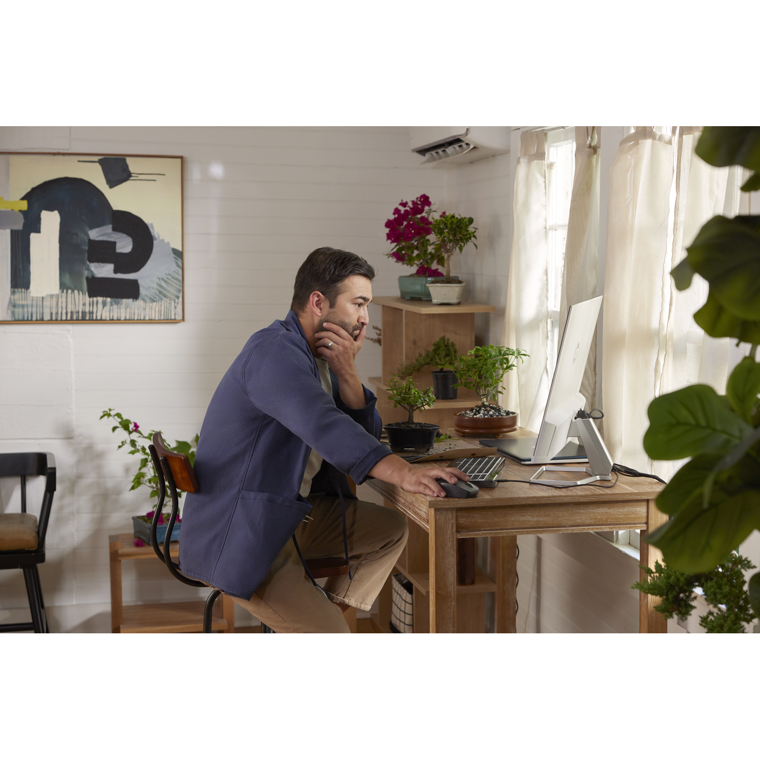 A man working at a desk, using a computer. Plants and a painting are in the background.