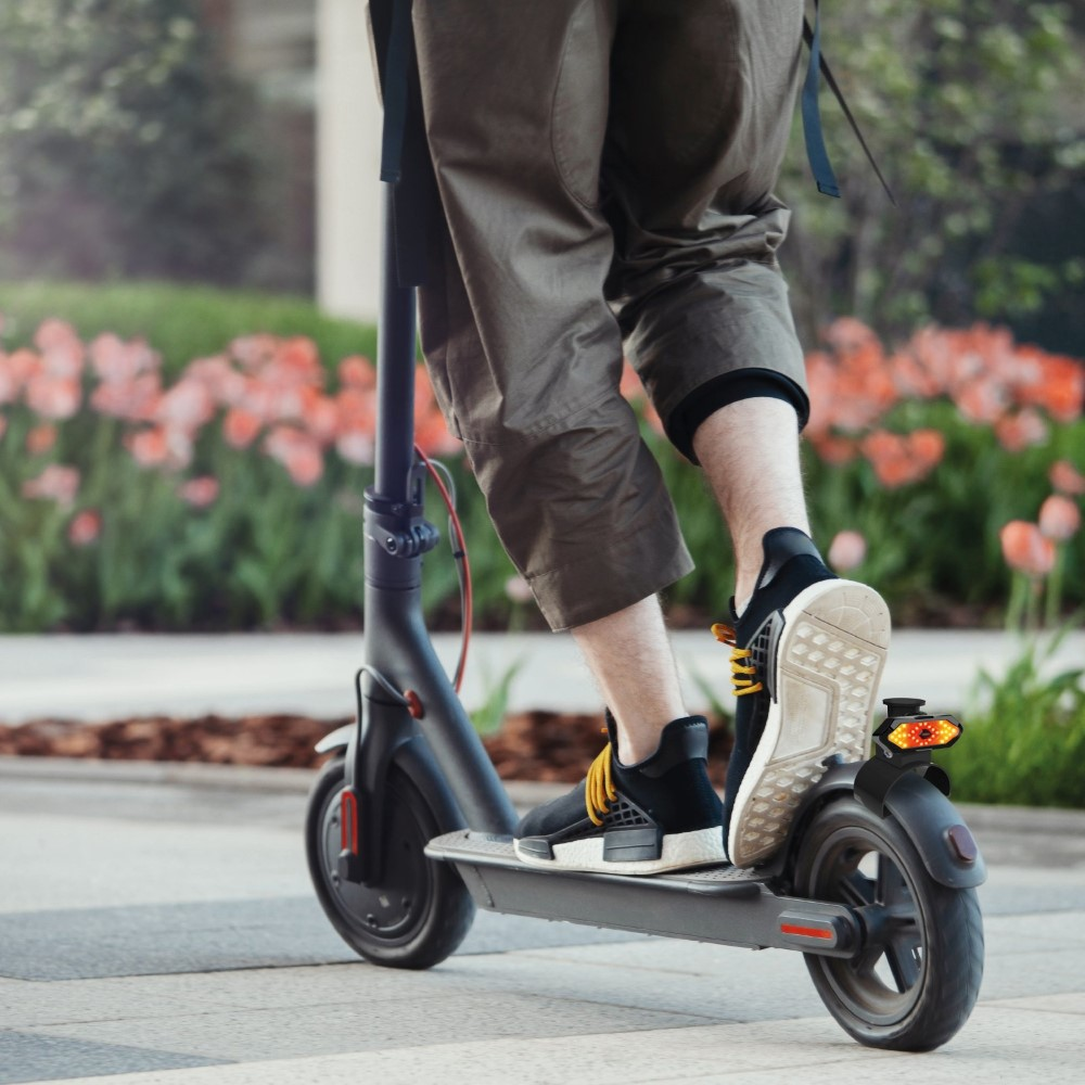 Osoba jedzie czarną hulajnogą elektryczną po utwardzonej ścieżce. Kwiaty w tle.